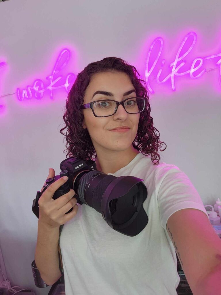 An selfie image of the photographer, Marisa Glaser, holding her camera and smiling. She is wearing a white t-shirt and standing in front of a neon pink sign that says "I Woke Up Like This"