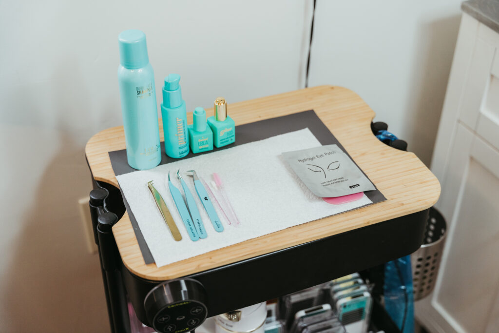 A wheeling cart with beauty service tools is set up. There are tweezers, eye patches, and blue bottles containing serums.