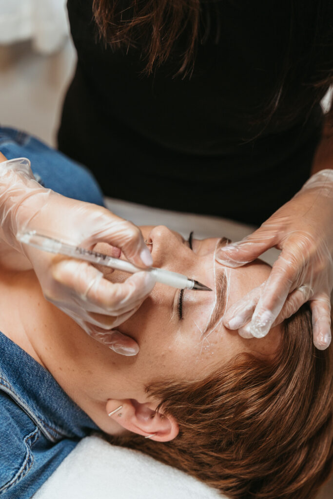 A woman uses a white pen to map out a new eye row shape on a client. A female client is laying down on the table below her, about to receive an eyebrow tint treatment.