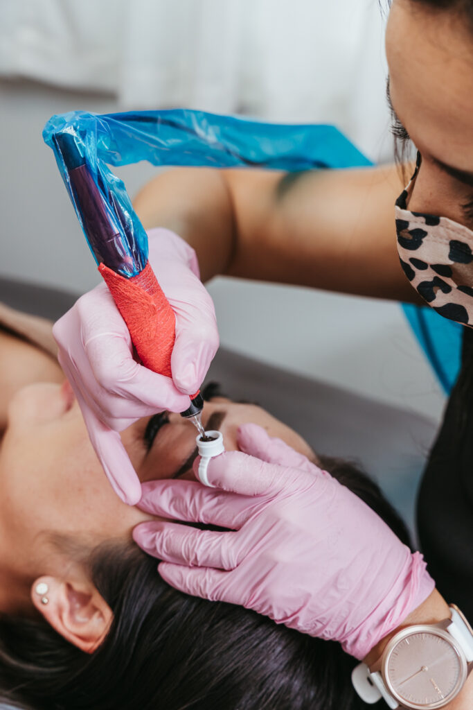 A woman wearing pink rubber gloves dips a tattoo machine into a pot of dark brown ink, about to add pigment to a client's eyebrows.