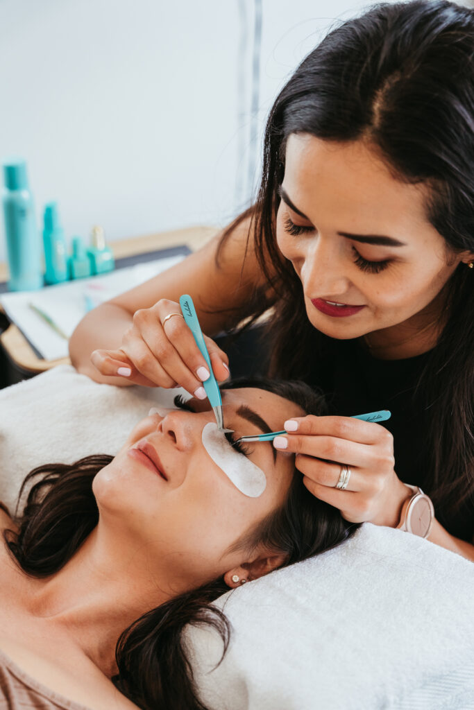 A woman holding an eyelash extension with a pair of blue tweezers. A female client with white eye patches on is laying down on the table below her, about to receive an eyelash treatment.