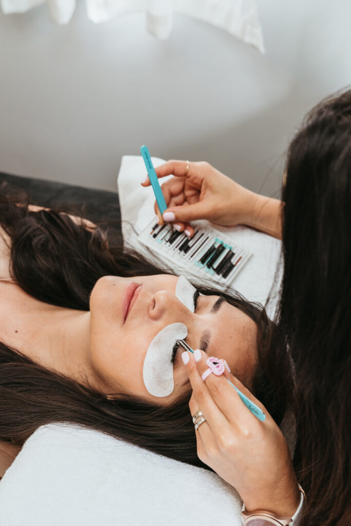 A woman holding an eyelash extension with a pair of blue tweezers. A female client with white eye patches on is laying down on the table below her, about to receive an eyelash treatment.