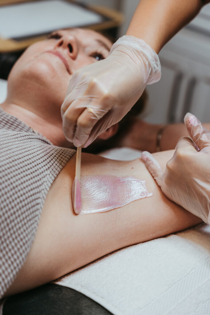 A female client laying on a table gets her armpit waxed by the esthetician wearing gloves.