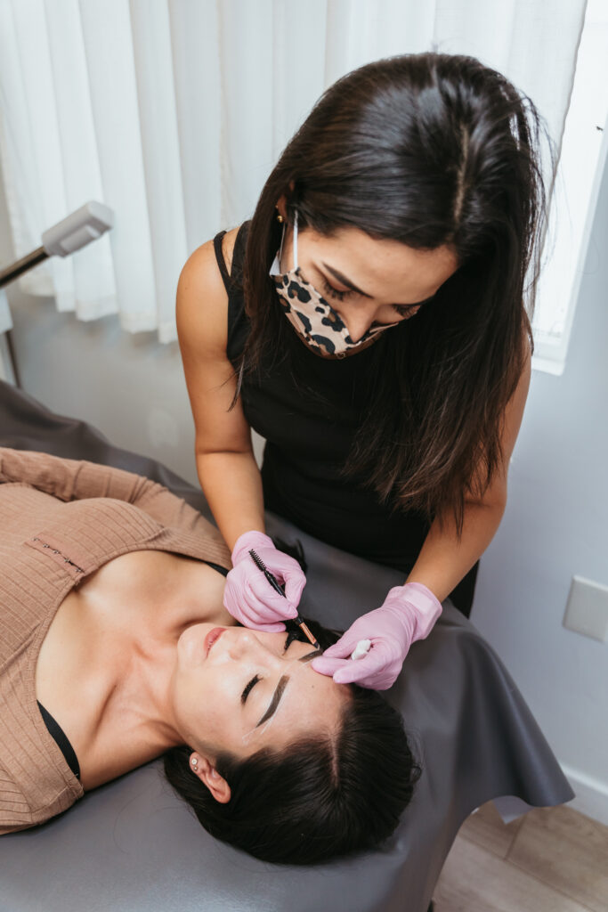 A woman wearing pink rubber gloves dips a brush into a pot of dark brown ink, about to add pigment to a client's eyebrows.