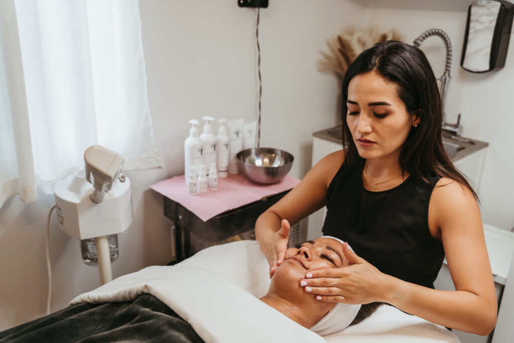 A female client laying on a table wrapped in a white towel and head wrap receives a facial massage.