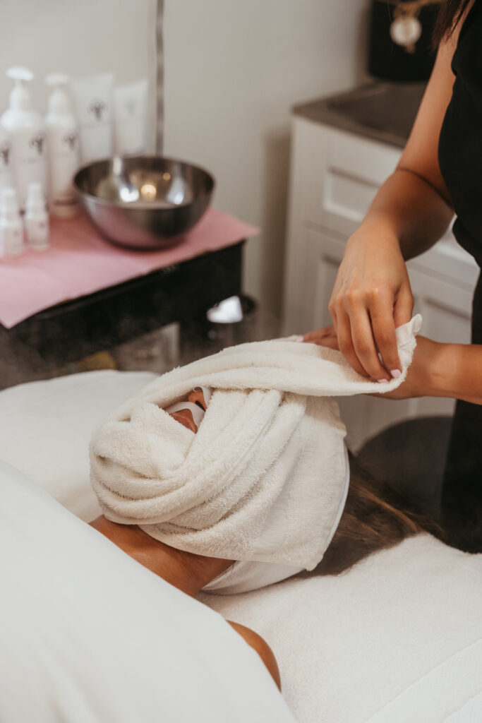 A female client laying on a table with a warm, damp, white towel being wrapped around her face prior to a massage.