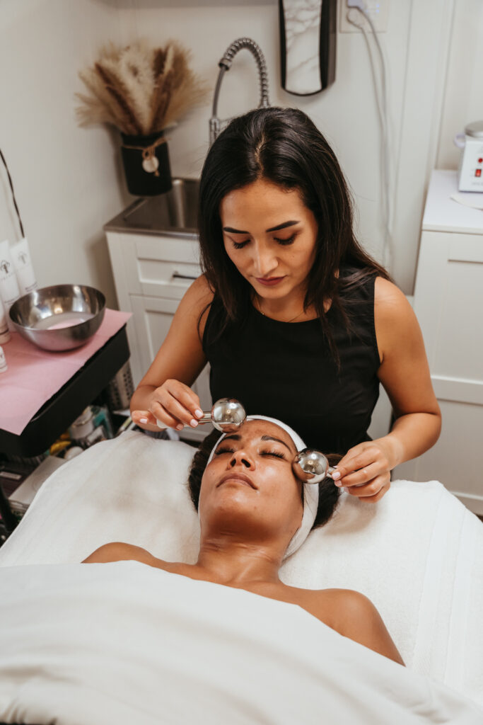 A female client laying on a table wrapped in a white towel and head wrap receives a facial massage using 2 metal ball-shaped tools.