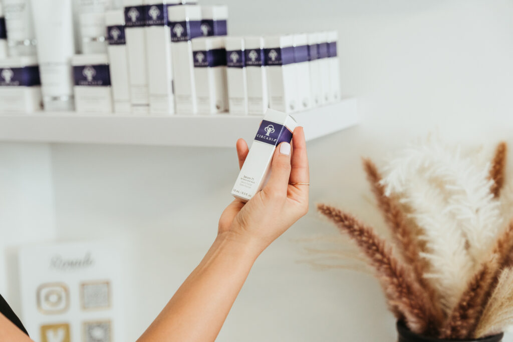 A woman holds a box of beauty product in front of a shelf. The box is white and dark blue and reads "CIRCADIA Serum"