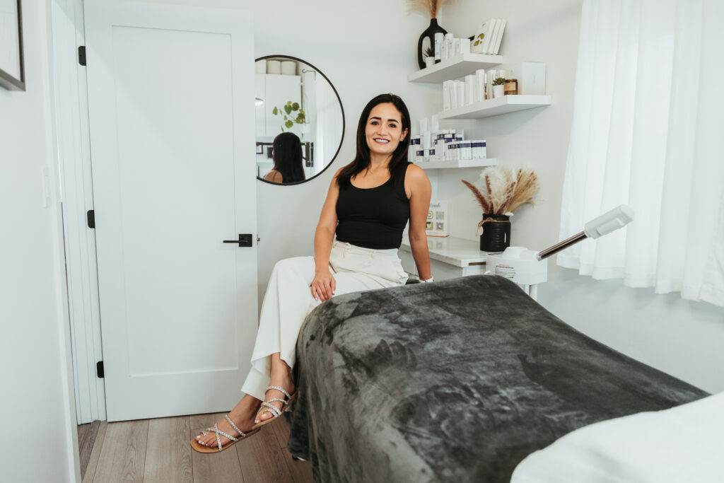 An esthetician sitting in her well-lit studio suite, posing for a headshot. She is wearing all black and smiling. A round mirror hangs on the wall behind her, reflecting part of the room's light décor.