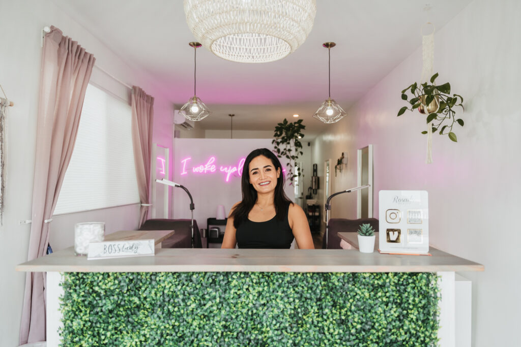 An esthetician standing at the front desk of her beauty salon, posing for a headshot. She is wearing all black and smiling. The room is well-lit and bright, with a pink neon sign that reads "I Woke Up Like This" in the background and plants hanging from the wall.