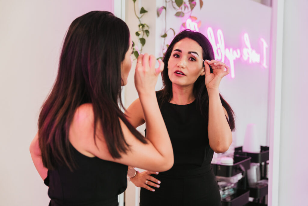 An esthetician looking in the mirror while holding a brush to her eyelashes. She is wearing all black and smiling. The room is well-lit and bright, with a pink neon sign that reads "I Woke Up Like This" in the background and plants hanging from the wall.