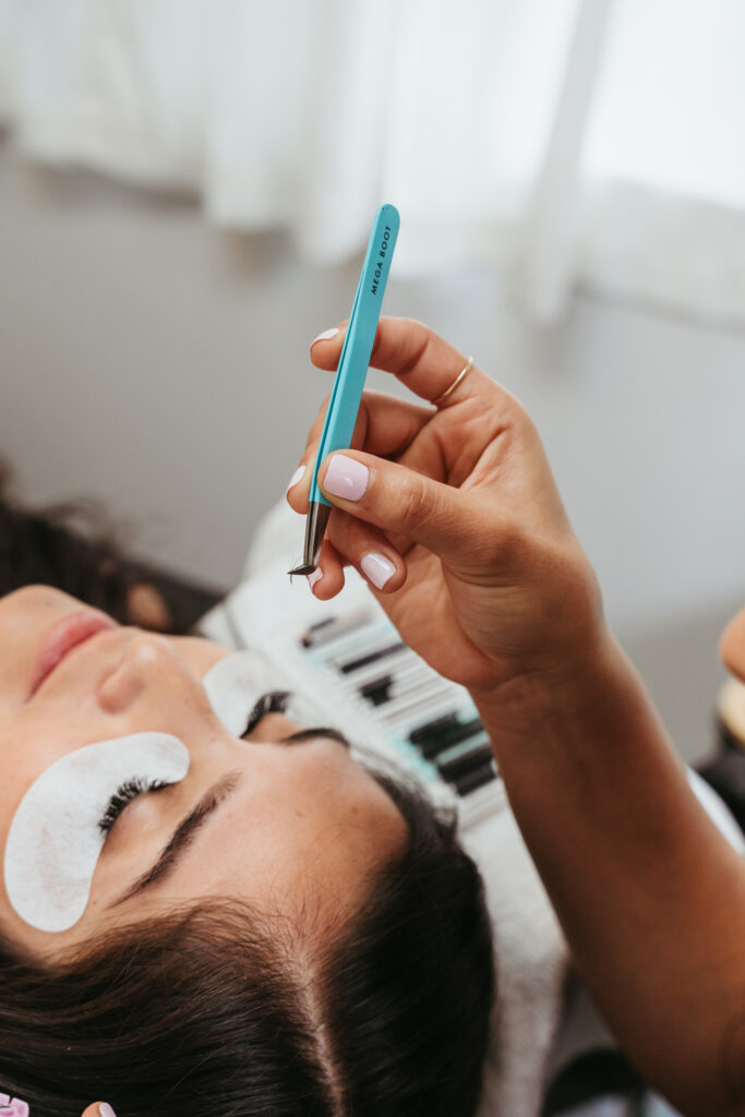 A woman holding an eyelash extension with a pair of blue tweezers. A female client with white eye patches on is laying down on the table below her, about to receive an eyelash treatment.