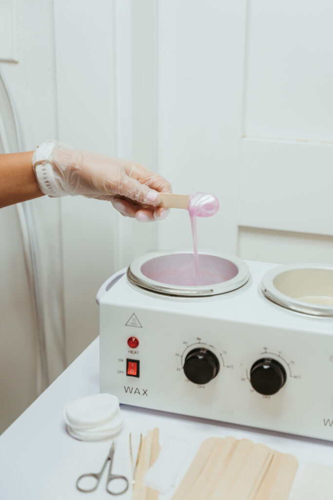 An image of a woman's hand wearing a clear rubber glove and dipping a popsicle stick into a white wax warmer filled with iridescent pink wax, to be used for facial or body waxing. 