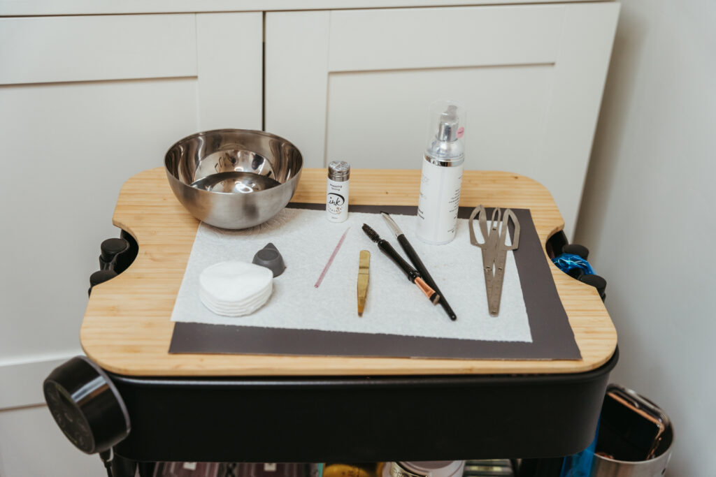 A wheeling cart with beauty service tools is set up. There are scissors, a metal bowl, cotton rounds, and a white bottle containing serum.