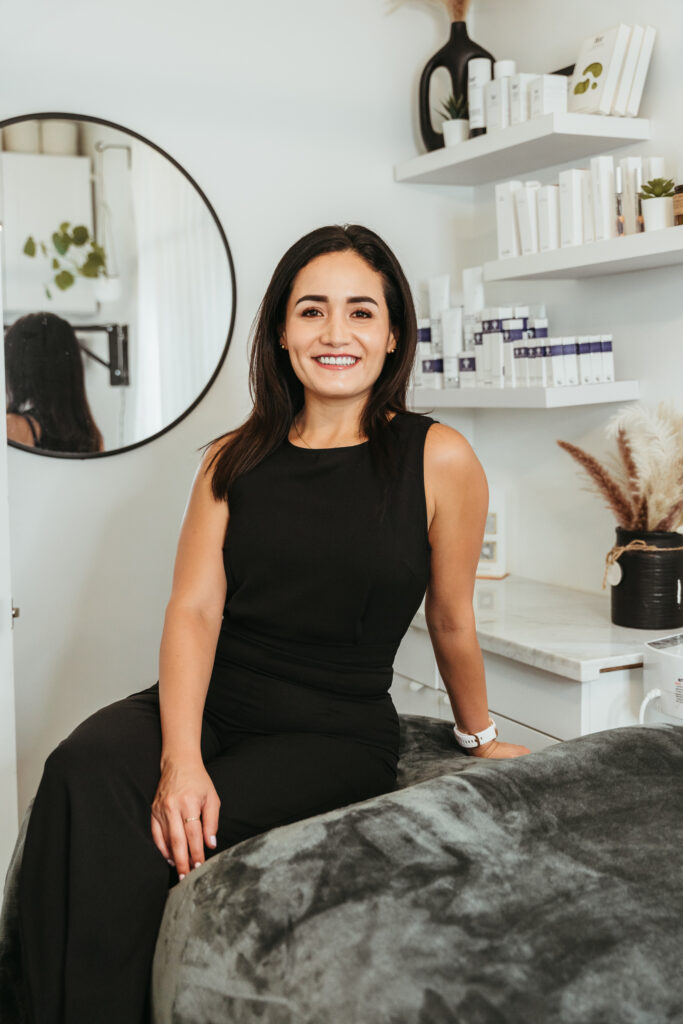 An esthetician sitting in her well-lit studio suite, posing for a headshot. She is wearing all black and smiling. A round mirror hangs on the wall behind her, reflecting part of the room's light décor.