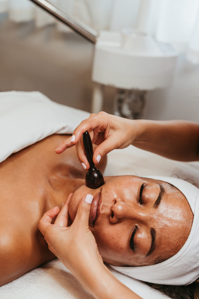 A female client laying on a table wrapped in a white towel and head wrap receives a facial massage using a black plastic tool.