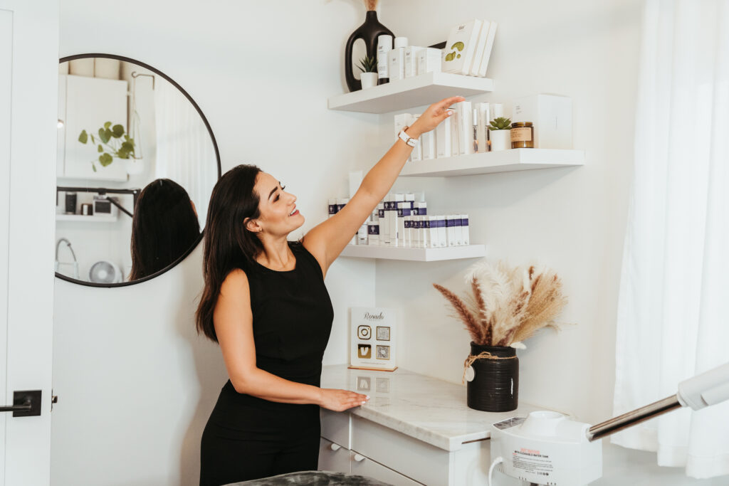 A woman dressed in black reaches for beauty products on her shelf in her salon suite.