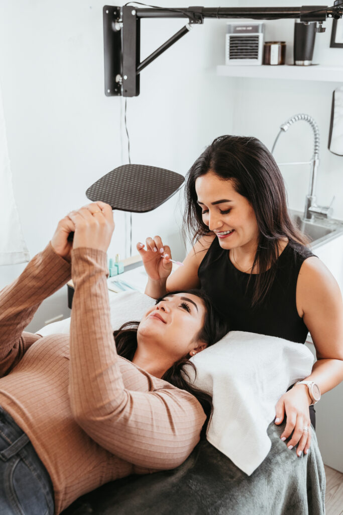 A woman is lying on a cushioned treatment bed while another woman stands beside her. The woman on the bed is holding a hand mirror up and smiling, while the woman next to her, in a black sleeveless top, appears to be adjusting or brushing the client’s eyebrows with a small tool. The room has a clean, bright aesthetic with a sink and shelves in the background.