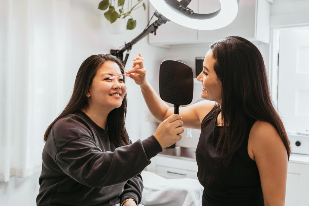 A woman seated on a stool is holding a hand mirror and smiling, while another woman, dressed in a sleeveless black outfit, gently applies a cosmetic tool to the seated woman's eyebrows. They are in a well-lit, cozy room, with plants hanging from the ceiling and a circular light overhead, adding a soft glow to the scene.