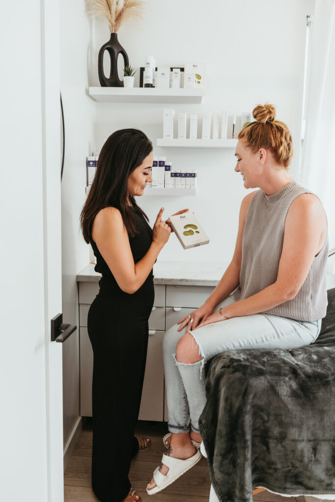 Two women are standing in a bright room having a friendly conversation. The woman on the left, wearing a black outfit, is showing a facial product to the woman on the right who is sitting and wearing a grey tank top and light colored jeans.