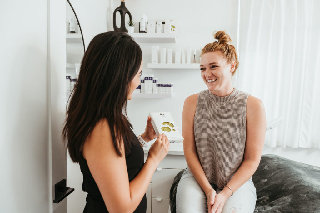 Two women are in a bright room having a friendly conversation about beauty tools. The woman on the left, wearing a black outfit, is showing a facial product to the woman on the right who is sitting and wearing a grey tank top and light colored jeans.