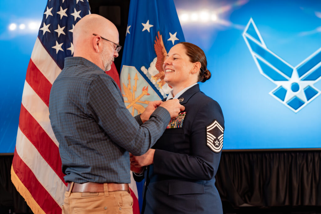 A woman in a U.S. military uniform, smiling with pride, receives a medal or pin from an older man during a formal ceremony. She wears a dress uniform with insignia on her sleeves, indicating a high rank. Behind them, a large American flag and a blue backdrop with military insignias add to the ceremonial atmosphere.