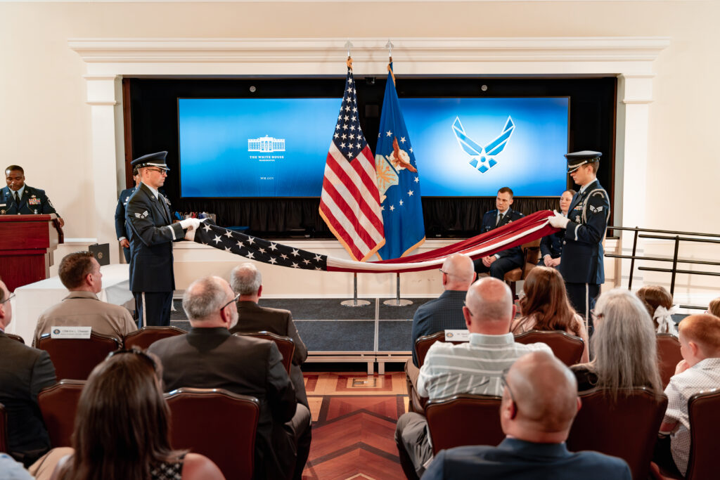 In a formal ceremony, service members in uniform stand on a stage about to fold an American flag. Audience members are seated, watching attentively. There are large television screens displaying military and White House emblems and flags in the background.