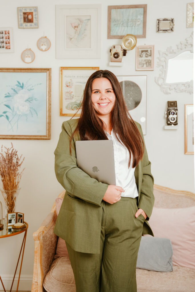 Woman in a business casual green outfit, standing and smiling with a laptop in hand, against a cozy, art-filled background.