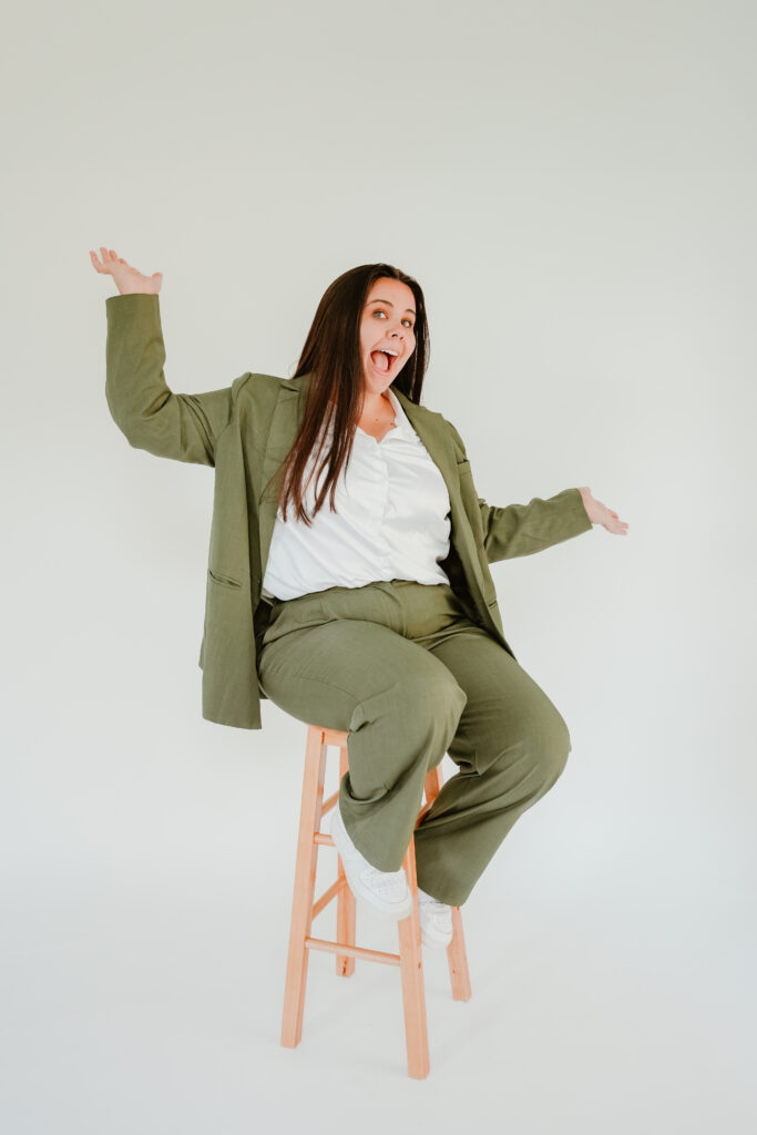 Energetic woman in a green suit raising her arms joyfully while seated on a wooden stool, white backdrop.