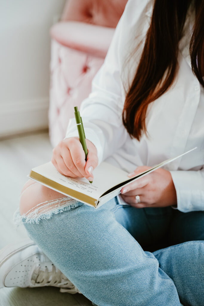 Detail shot of a woman writing with a green pen in a notebook, with blue jeans and white shirt visible.