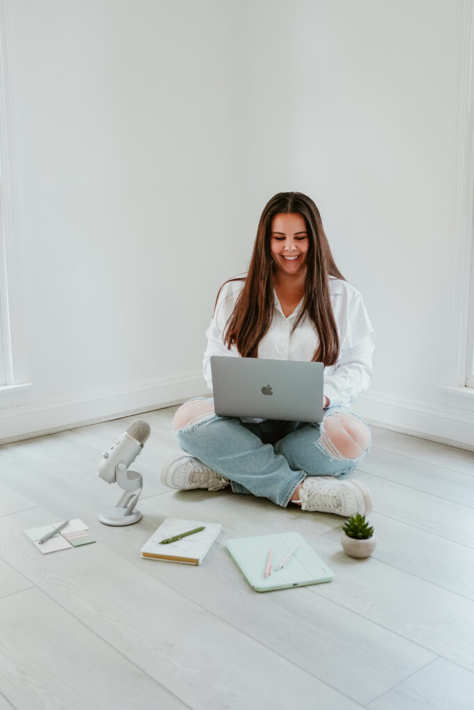 Casual workspace setup with a woman sitting on the floor using a laptop, with items like a notebook, a podcast microphone, and plants around her.