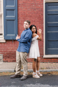 Man and woman posed back to back doing a "Charlie's Angels" pose in front of a red brick wall.