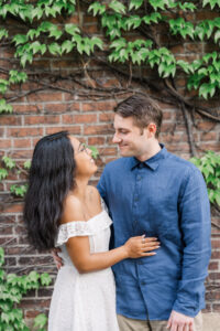 The couple smiling at each other in front of a brick wall with ivy.