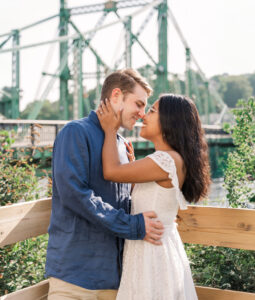 Romantic portrait of the engaged couple embracing by a historic suspension bridge in soft morning light