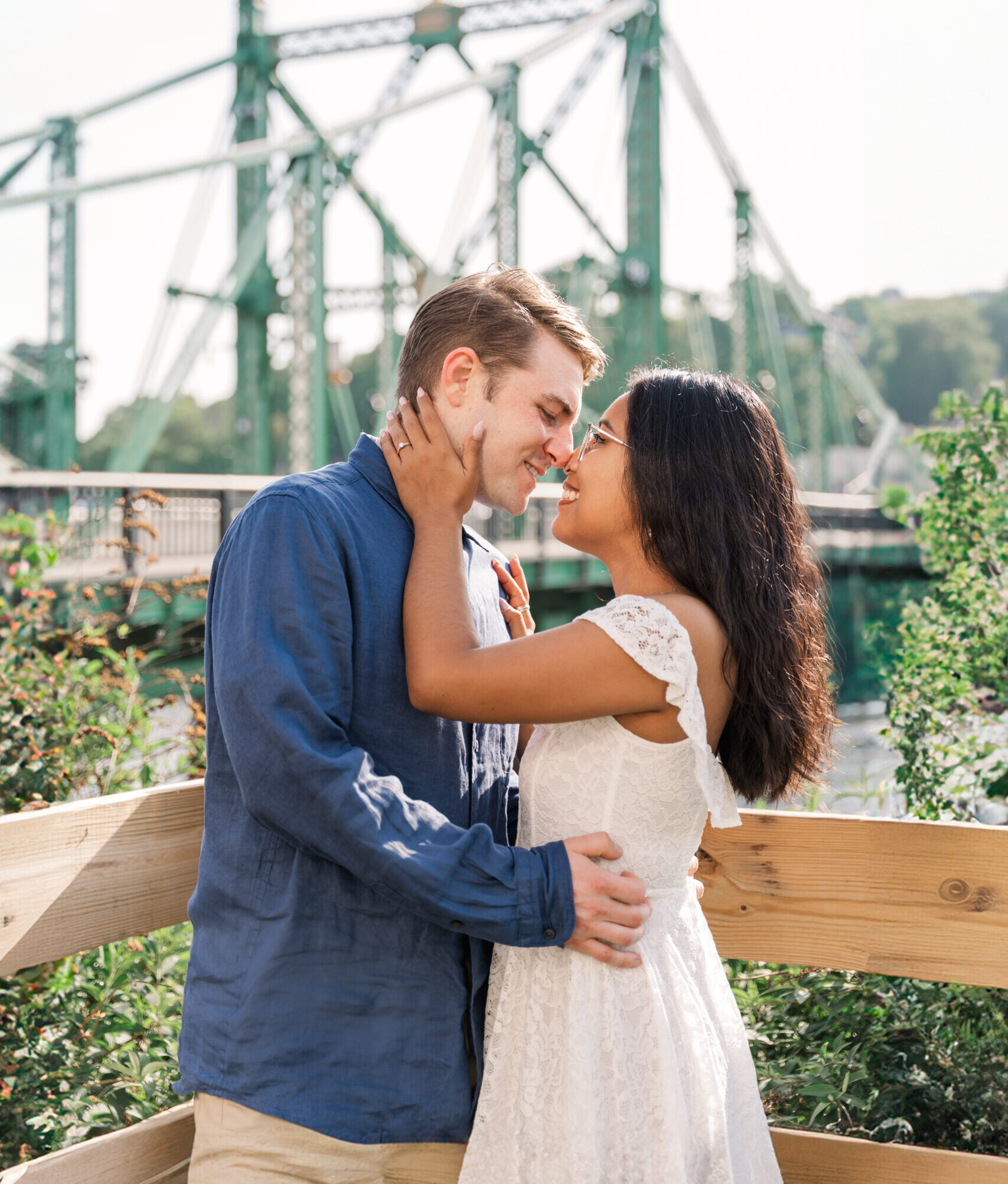 Romantic portrait of the engaged couple embracing by a historic suspension bridge in soft morning light