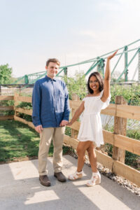 Woman holding her fiancé's hand and posing with her other hand up, while the man makes a skeptical expression.