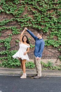 A joyful couple shares a spin during their engagement session, with the woman in a white dress and the man in a blue shirt and khakis.