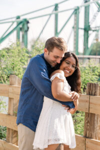 The man hugs his fiancée from behind, both smiling brightly near a scenic walkway.