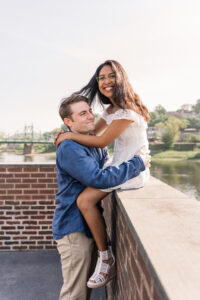 The woman laughs as her fiancé gives her a lift with the water and greenery behind them.