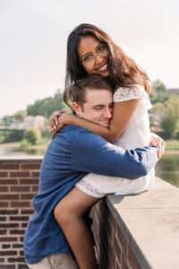 The woman smiles as her fiancé hugs her with the water and greenery behind them.