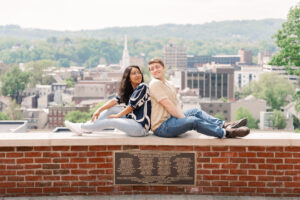 The couple poses back-to-back on a brick ledge with a panoramic view of downtown buildings and church steeples.