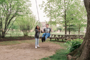 A woman stands beside a man sitting on a swing. They're both in a wooded park area with picnic tables, smiling and wearing matching themed shirts.
