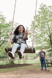 A woman smiles and shows off a ring while swinging forward on a wooden swing. She's mid-air and wearing a "feral era" shirt, with trees and a man in the background.