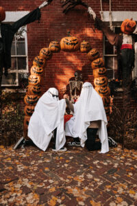 Two people in ghost costumes hold hands while kneeling in a playful pose under an arch of jack-o-lanterns.