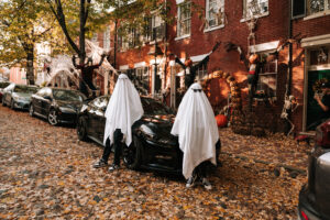 A ghost-costumed couple sits back to back on the hood of a car, draped in white sheets and sunglasses with leaves all over the street..