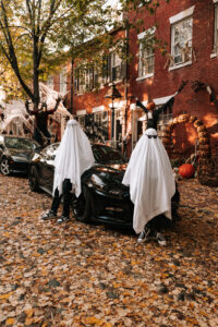 A couple wearing ghost costumes with sunglasses leans playfully against a vintage car.