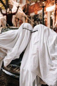 A couple in ghost costumes nudge their noses together against a car hood.