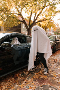 Two ghost-costumed people stand in the middle of a cobblestone street lined with parked cars and autumn leaves, one is driving a car.