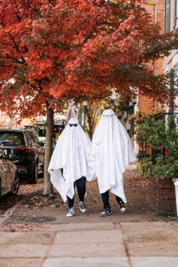 Two people in ghost sheets and sneakers pose on a leaf-covered sidewalk with fall trees behind them.
