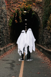 A ghost-costumed couple sits back to back in front of the entrance to a historic tunnel covered in ivy.